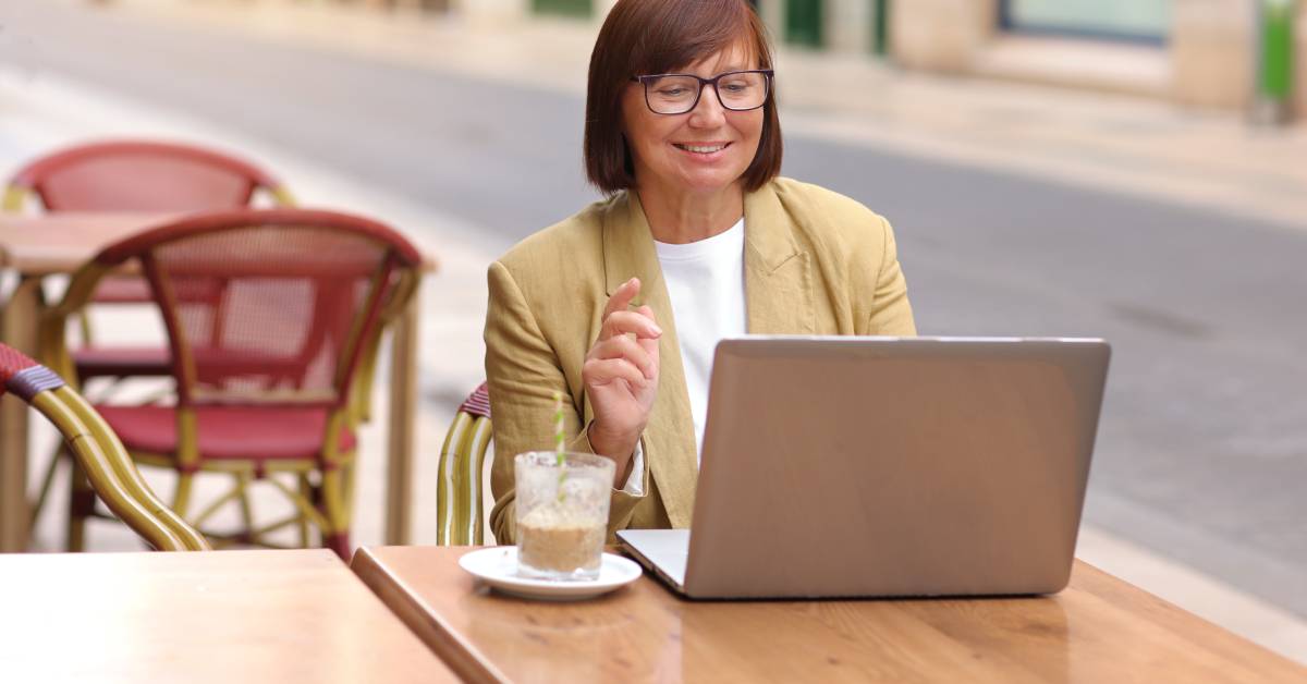 A person wearing glasses is sitting at a brown square table outside and smiling while using their laptop.