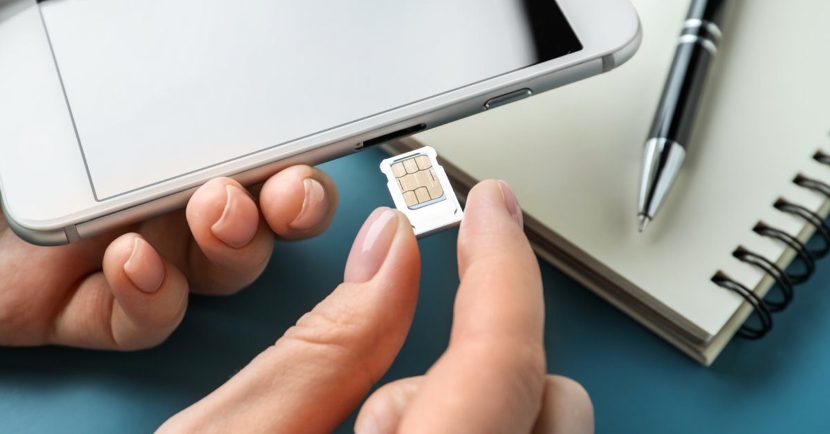 A hand is inserting a small white SIM card into an opening on the side of a large gray cellphone with a clean screen.