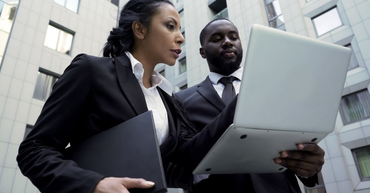 Two people dressed in business suits are outside and looking at a laptop. They are standing next to a gray building.