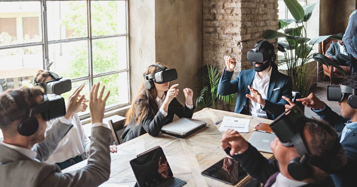Six employees are wearing VR headsets while sitting around a large wood table with tablets and laptops in front of them.