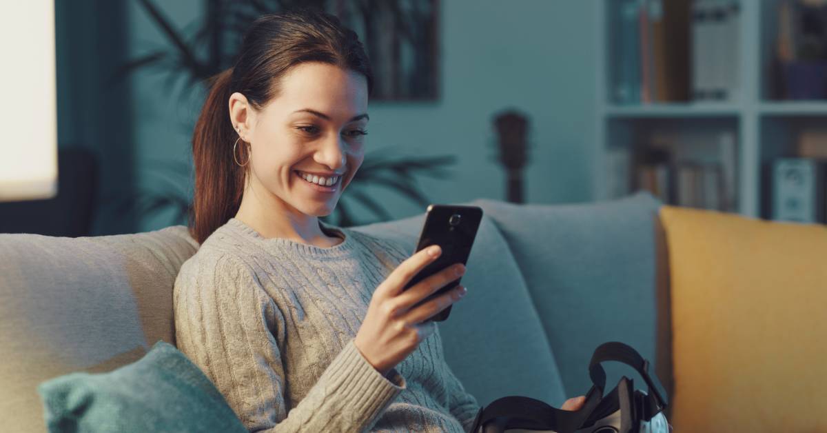 A person wearing a sweater sits on a couch and smiles while looking at their cell phone. A VR headset is in their lap.
