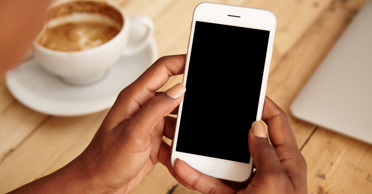 A person sits at a wooden table. A mug of coffee is on the table and the person holds a cellphone with a blank screen.