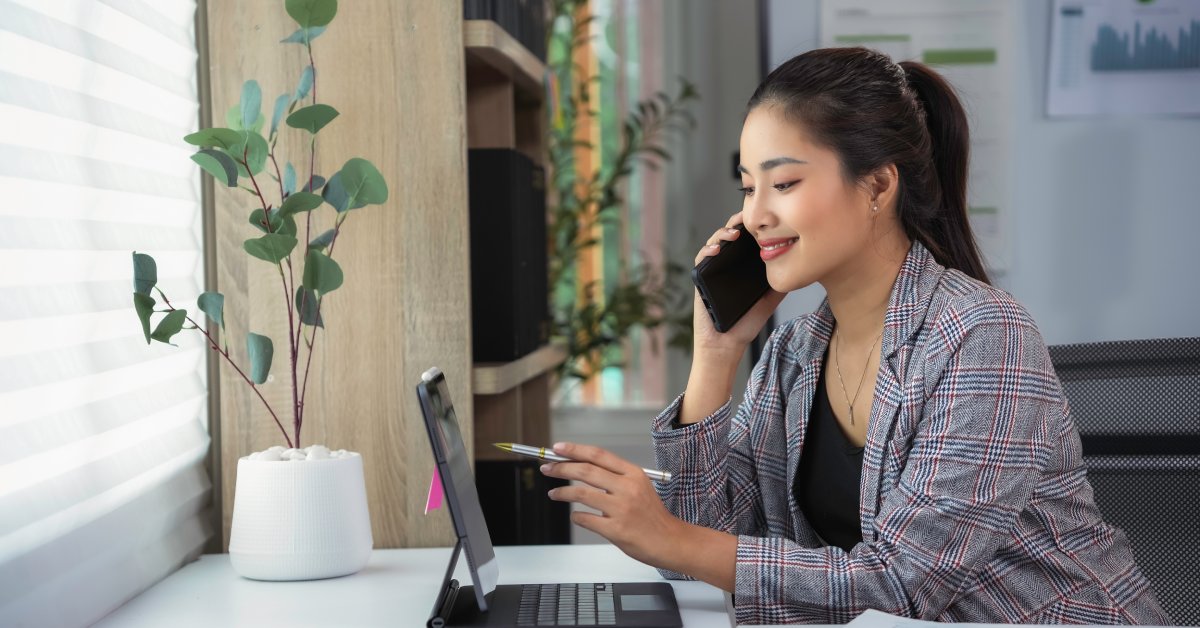 An employee wearing a suit is talking on the phone while sitting at a desk and pointing at a laptop with their pen.