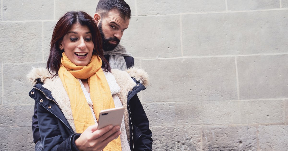A person wearing a yellow scarf looks at their cellphone. Another person stands behind them and looks at the phone.