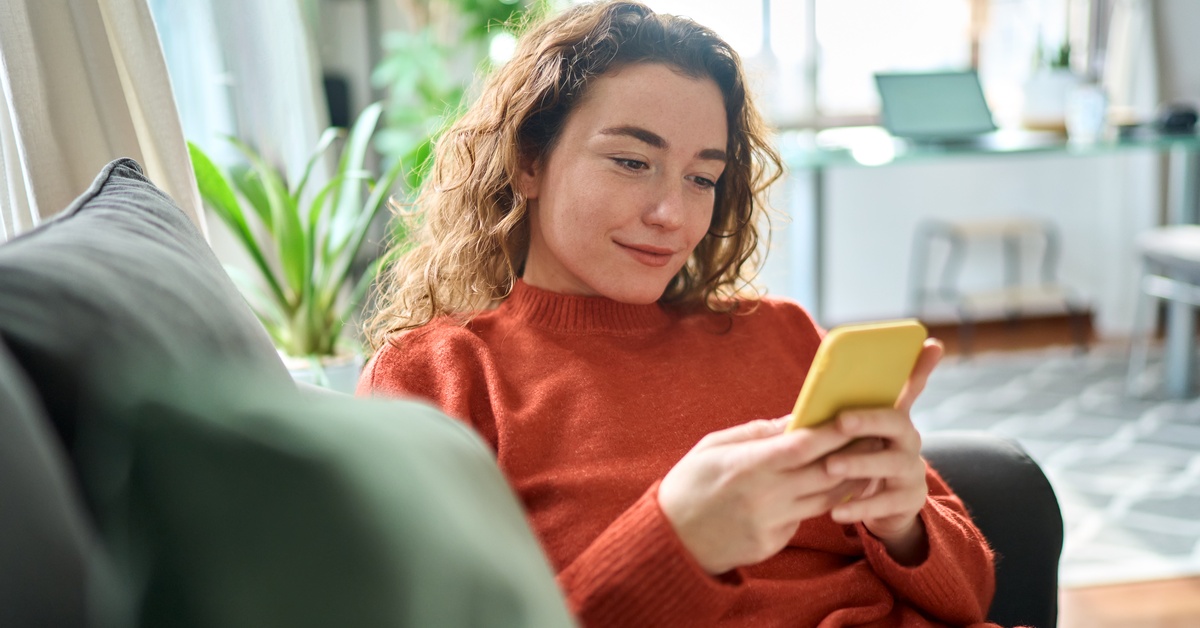 A person wearing an orange sweatshirt sits on a couch. They are holding a yellow cell phone in their hands.