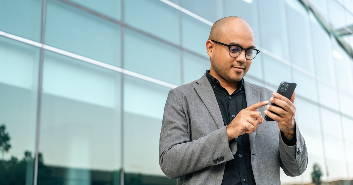 A person wearing a business suit and glasses stands outside near windows and holds a cell phone in their hands.