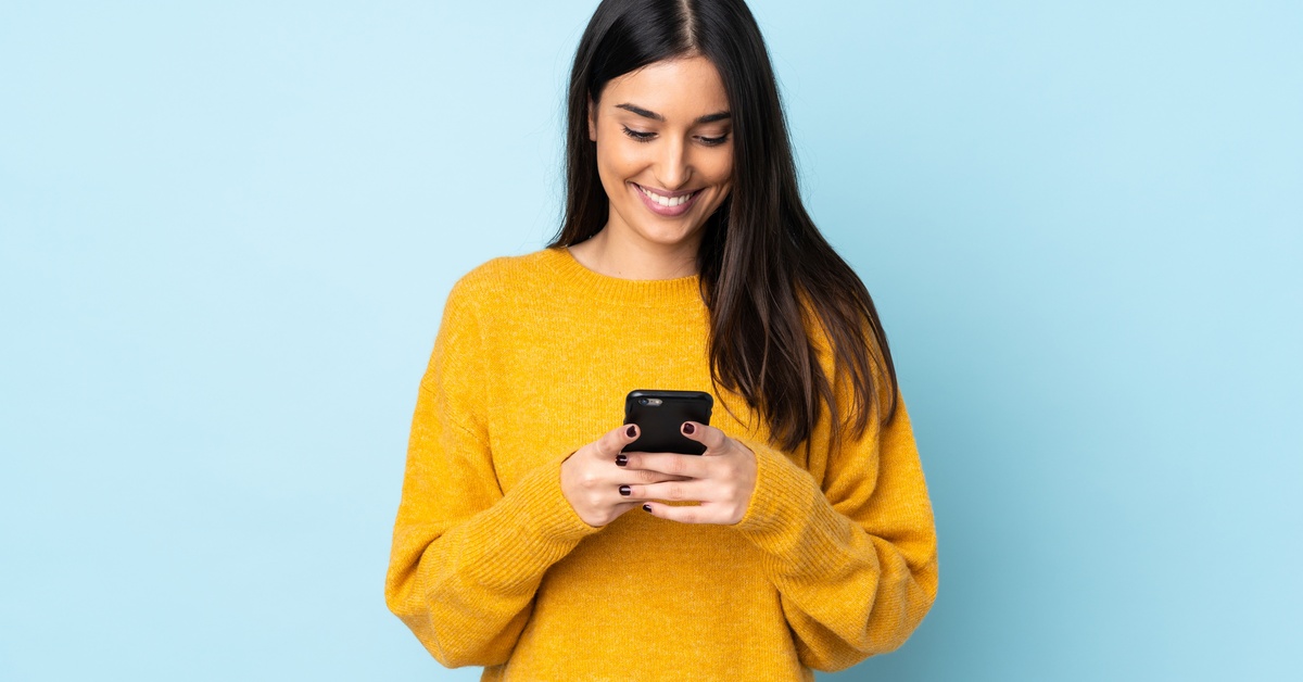 A person wearing a yellow sweater stands in front of a light blue wall while looking down at their cell phone.