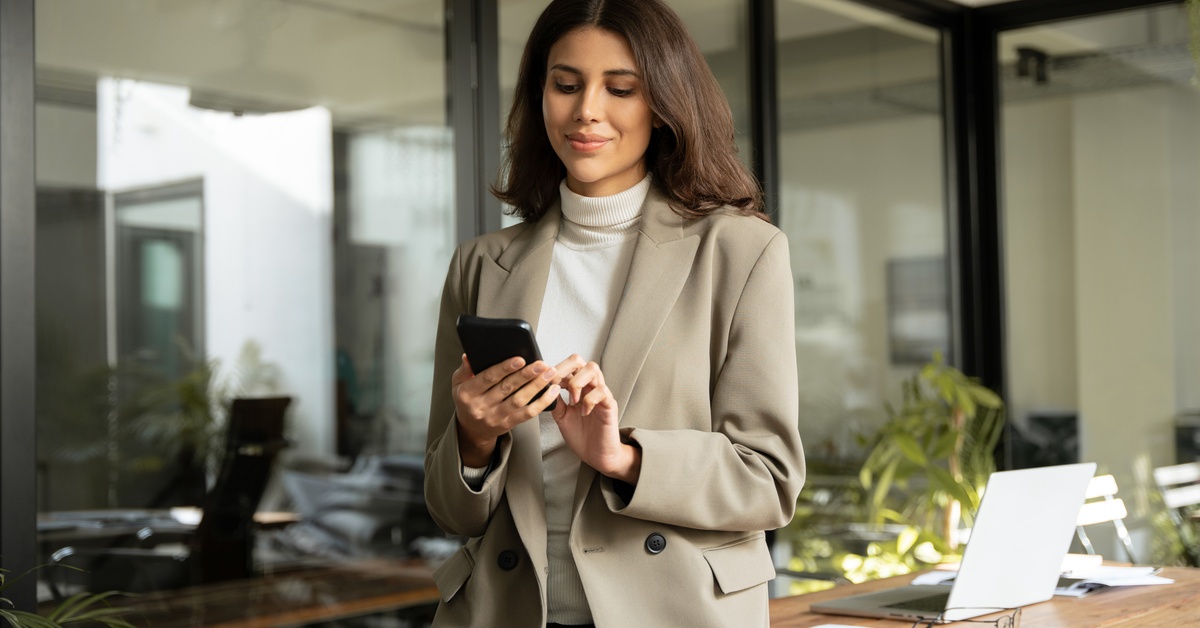 A person wearing a gray jacket stands in an office and swipes the screen of a small cellphone that is in their hands.