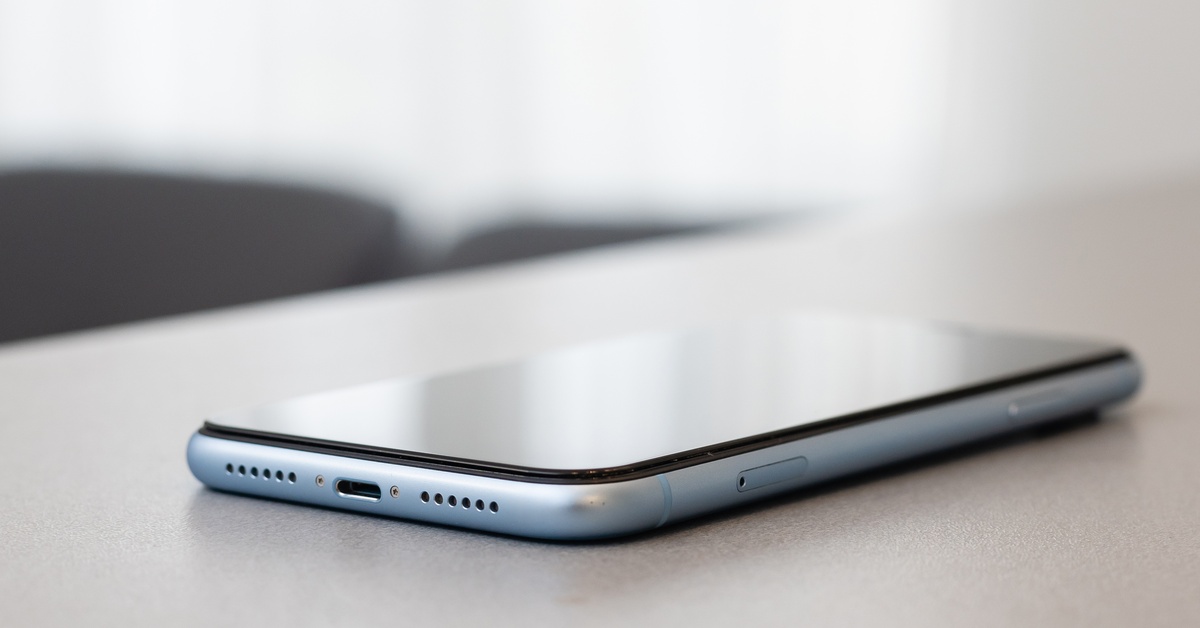 A small cellphone lying down on a gray table. The phone screen is facing the ceiling and reflecting a nearby window.