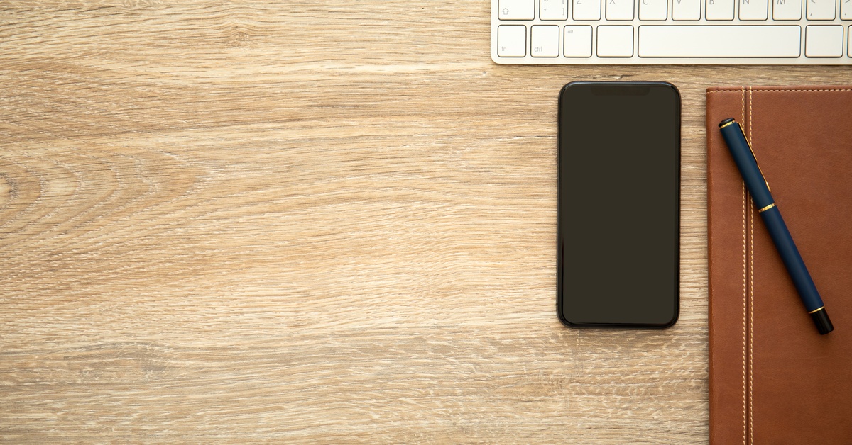 A black cellphone, a brown notebook, and a white keyboard sit on a wood surface. A pen is on top of the notebook.