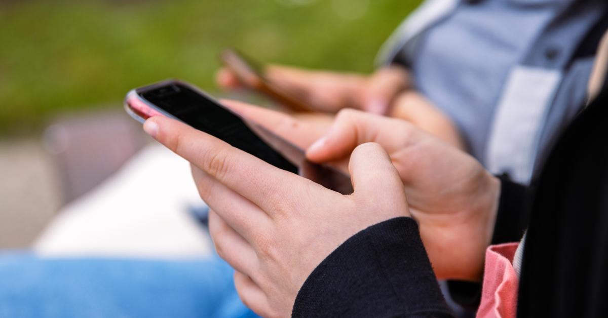 Two people are sitting outside during the daytime. They are next to green bushes and using their cell phones.