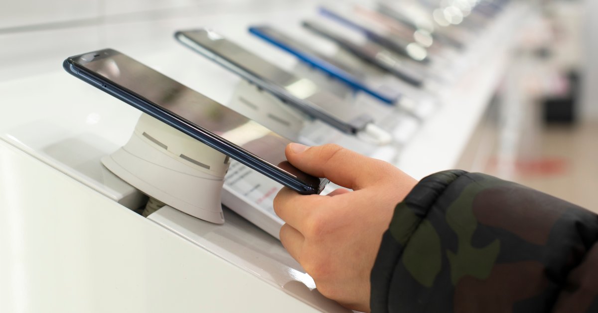 A person wearing a camouflage jacket is placing their left hand on a cell phone in a display at a phone store.