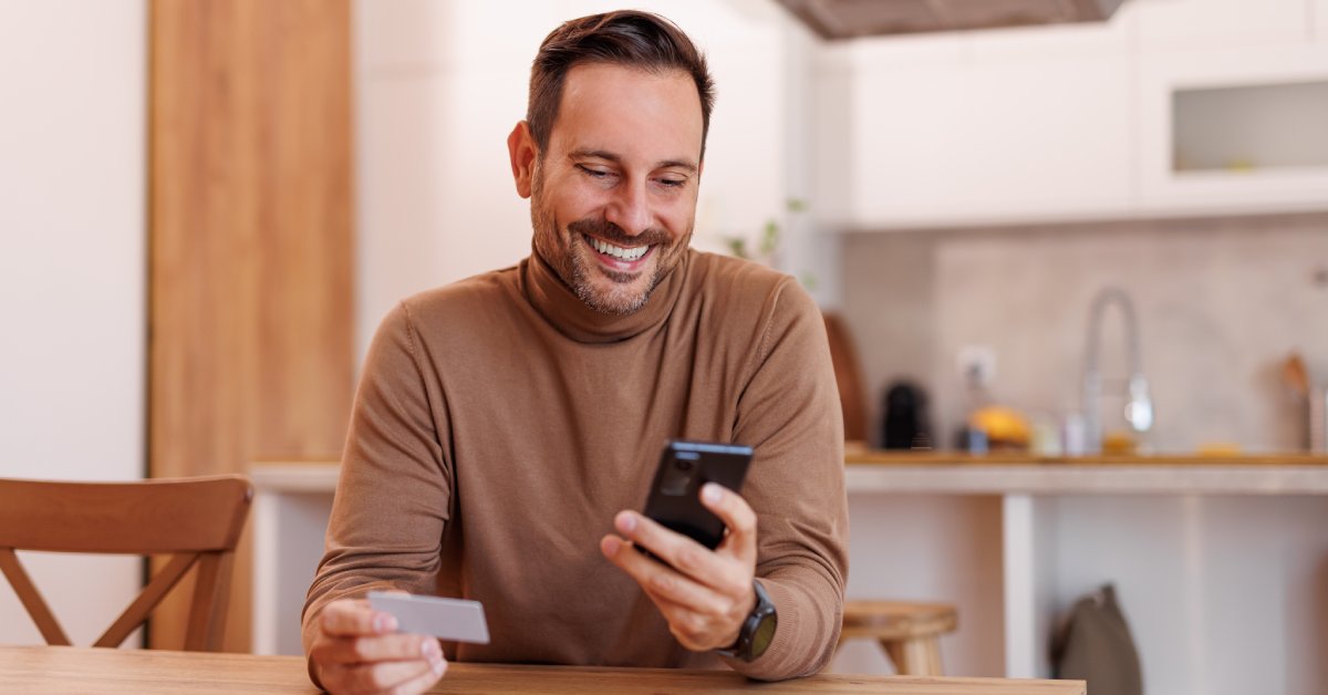A person wearing a brown sweater smiles while holding a cell phone in their left hand and a card in their right hand.