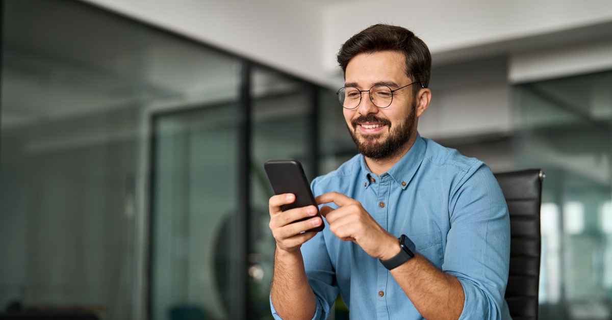 A person wearing a blue shirt is smiling and scrolling on their cell phone while sitting in an office.