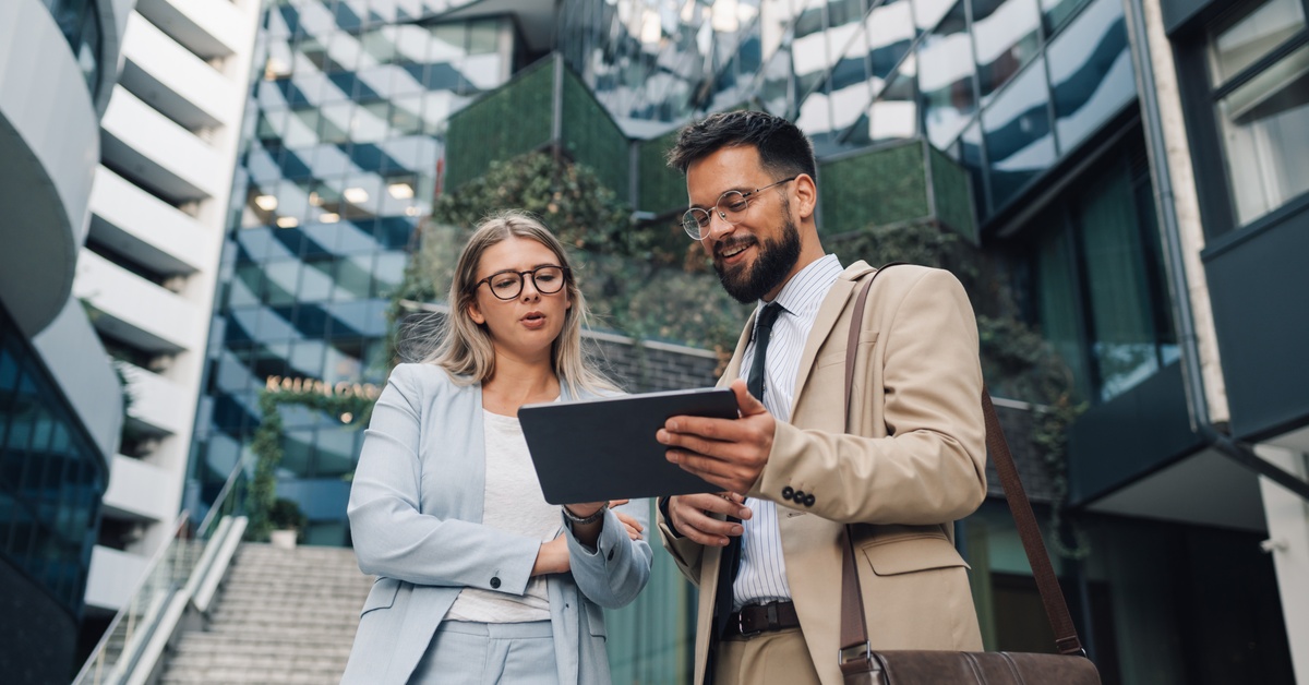 Two professionally dressed people standing outside in front of a skyscraper. They are both looking at a tablet and talking.