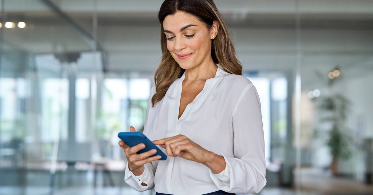 A smiling woman holding and using a blue smartphone in the middle of a brightly lit office space with glass partitions.