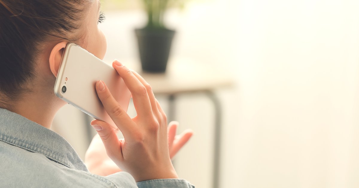 A woman wearing a jean jacket is sitting and holding her cell phone up to her ear. Her other hand is moving in a gesture.