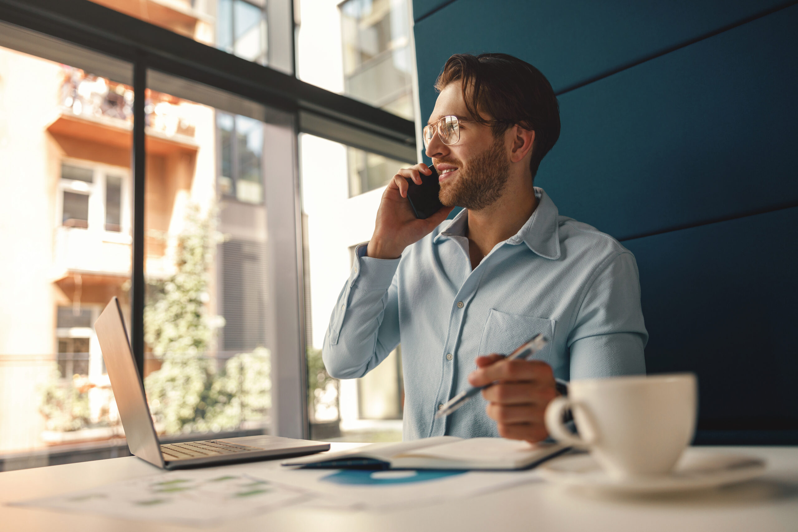 A businessman sits at a desk next to a window. He is on the phone and hold a pen above a notebook in the other hand.