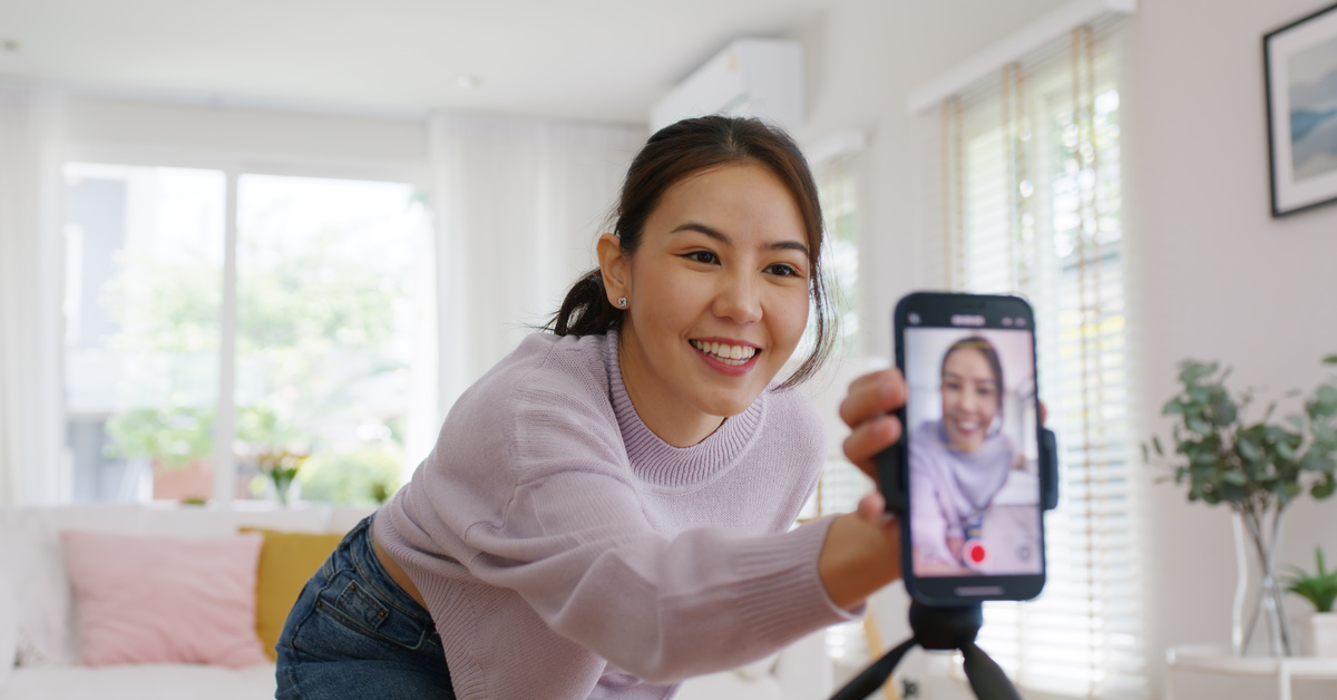 A woman crouches down in front of a tripod with a phone. Her hand is on the phone and she smiles through the camera.