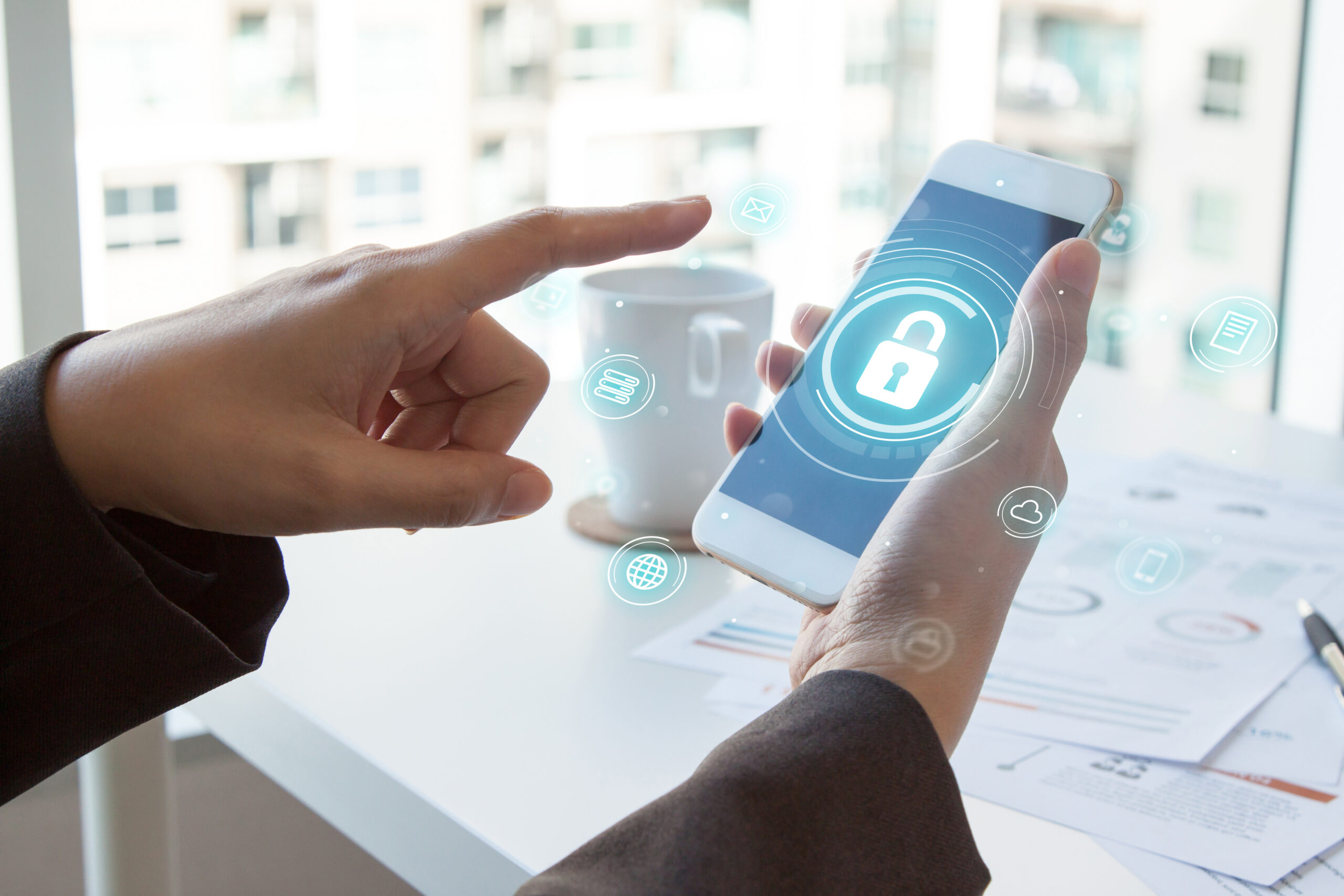 A person holds a cell phone up and points with one finger at the screen. Various security icons hover over the phone.