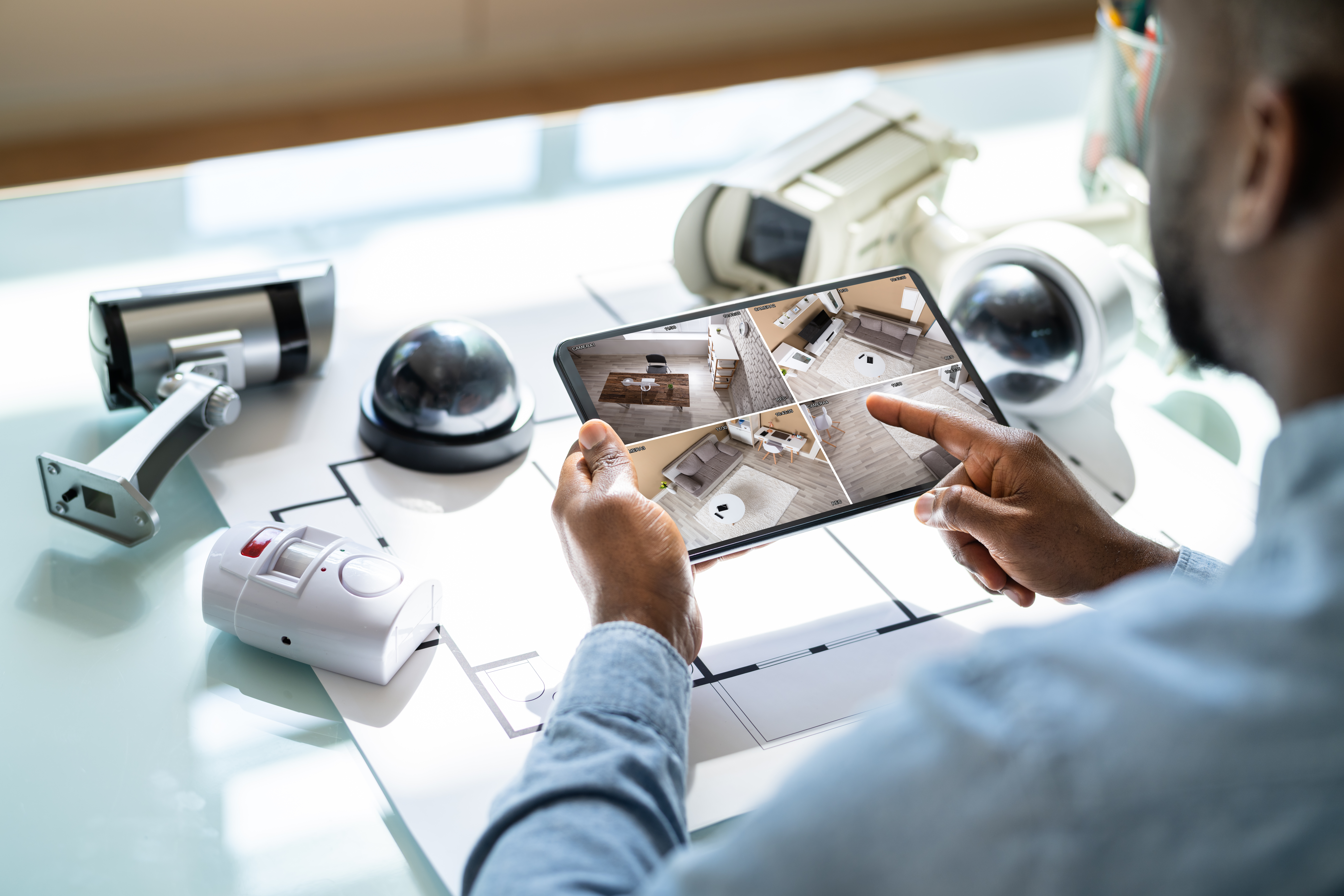 A man sits at a desk and holds a tablet with home security footage. Various security cameras are lying on the desk.