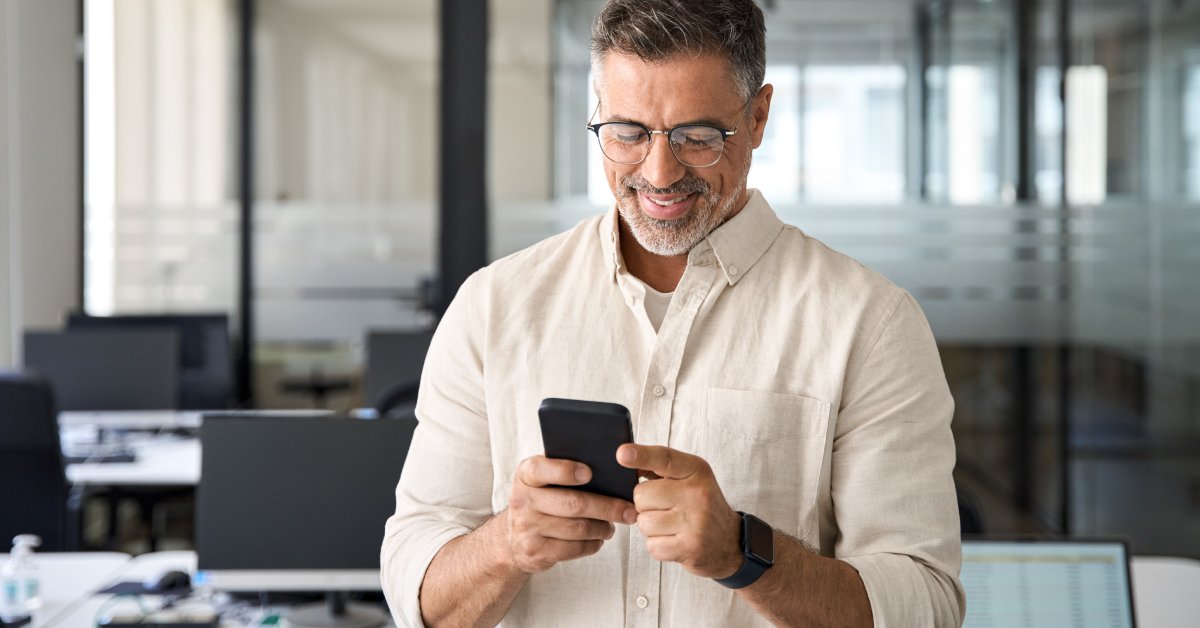 A man wearing a white shirt and glasses stands in an office while holding a phone in his hands and smiling at the screen.