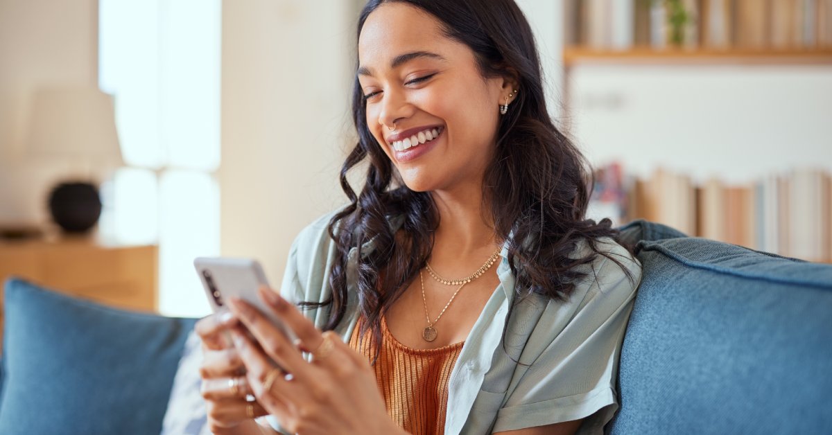 A woman holds a phone and smiles at it while sitting on a blue couch with a bookshelf and lamp in the background.