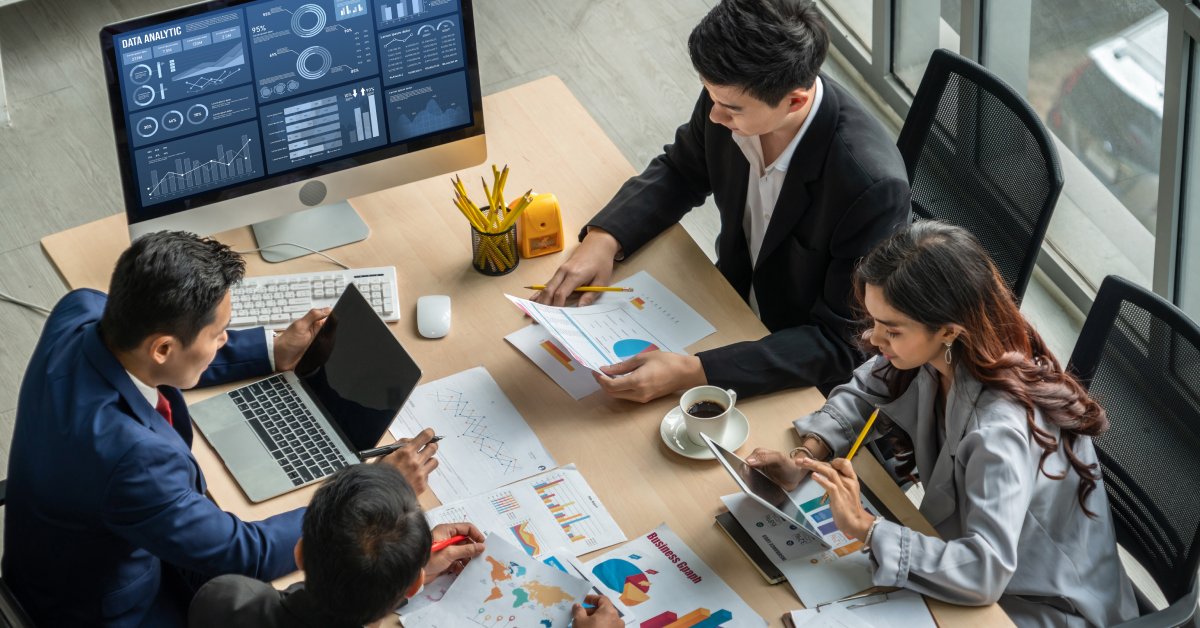 A group of business professionals sitting around a desk. Data, graphs, computers, and tablets are in front of everyone.