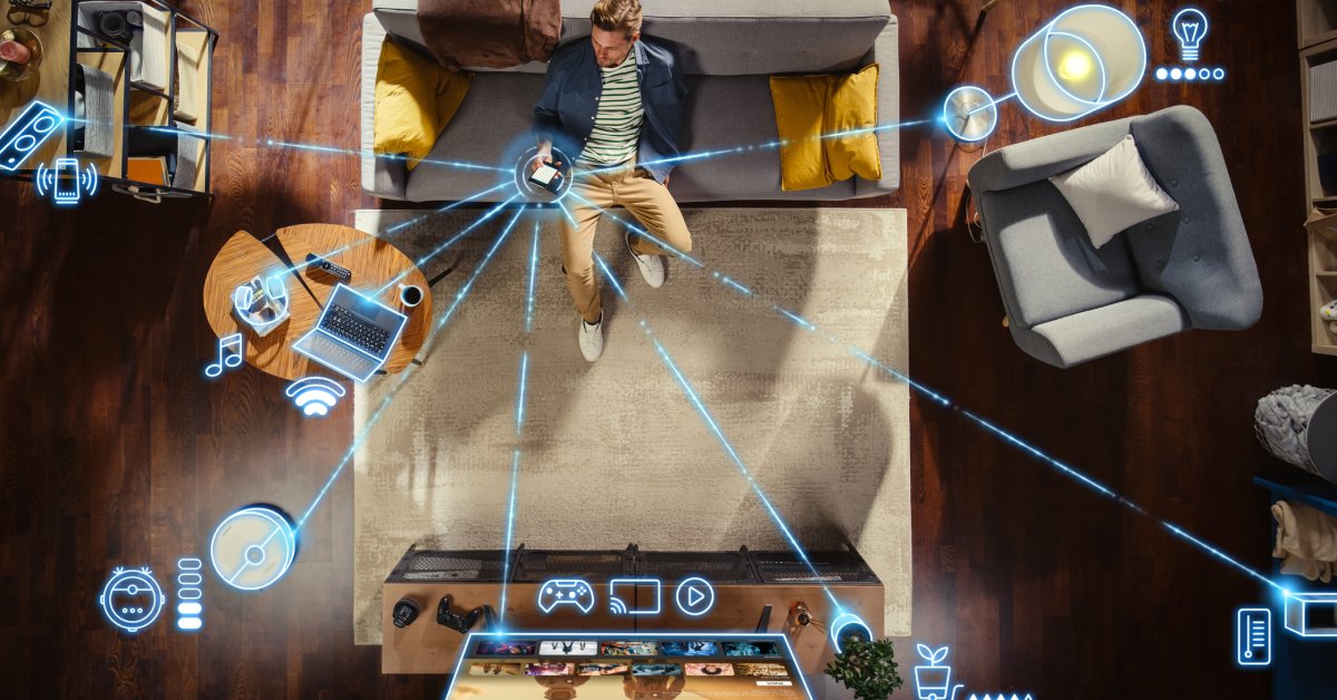 A man is sitting on his couch and holding his phone. Blue lines and graphics show his phone connected to numerous other devices.