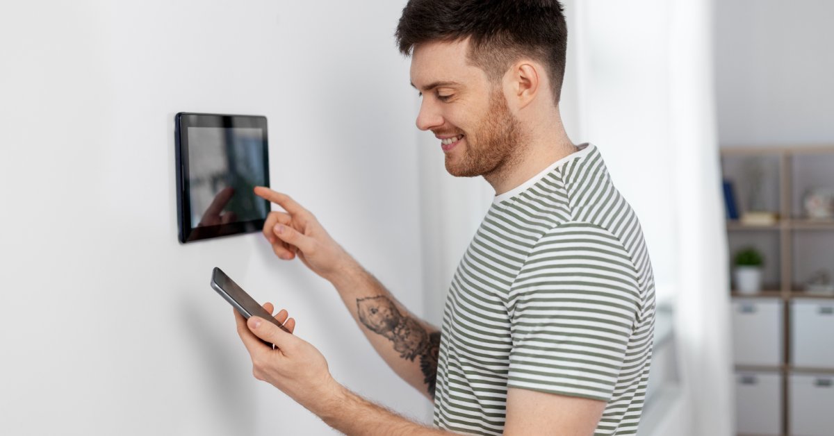 A man stands in front of a tablet on the wall of his home. He holds his phone in the other hand and smiles at the screen.