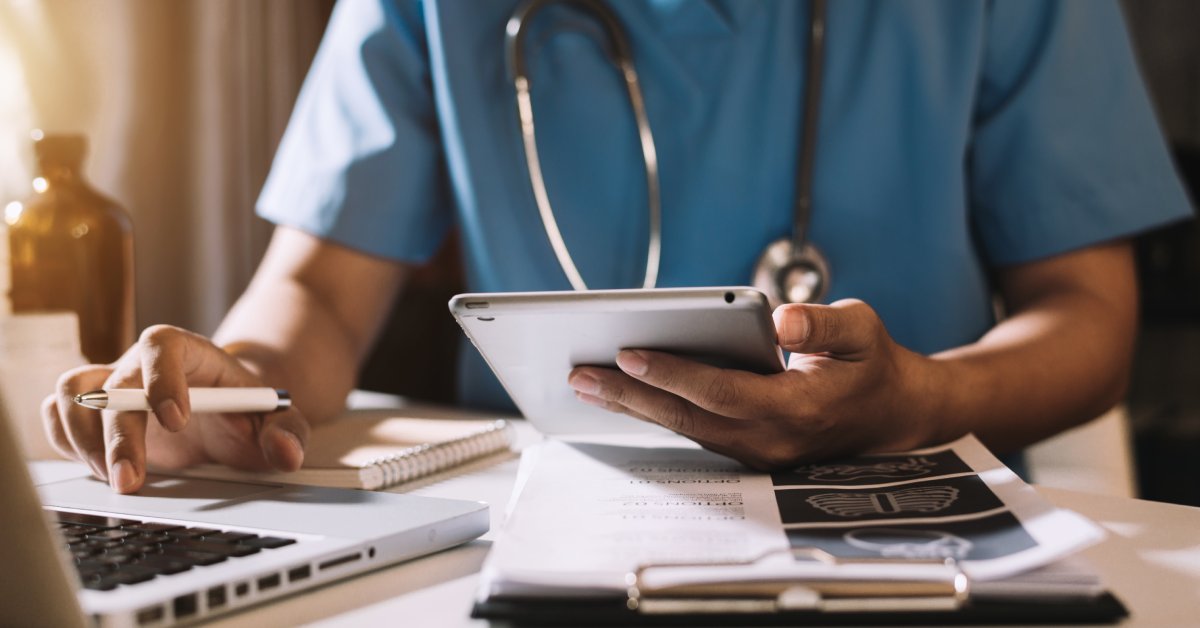 Someone wearing scrubs and a stethoscope sits at a desk. They hold a tablet and pen in their hands with X-rays on the desk.