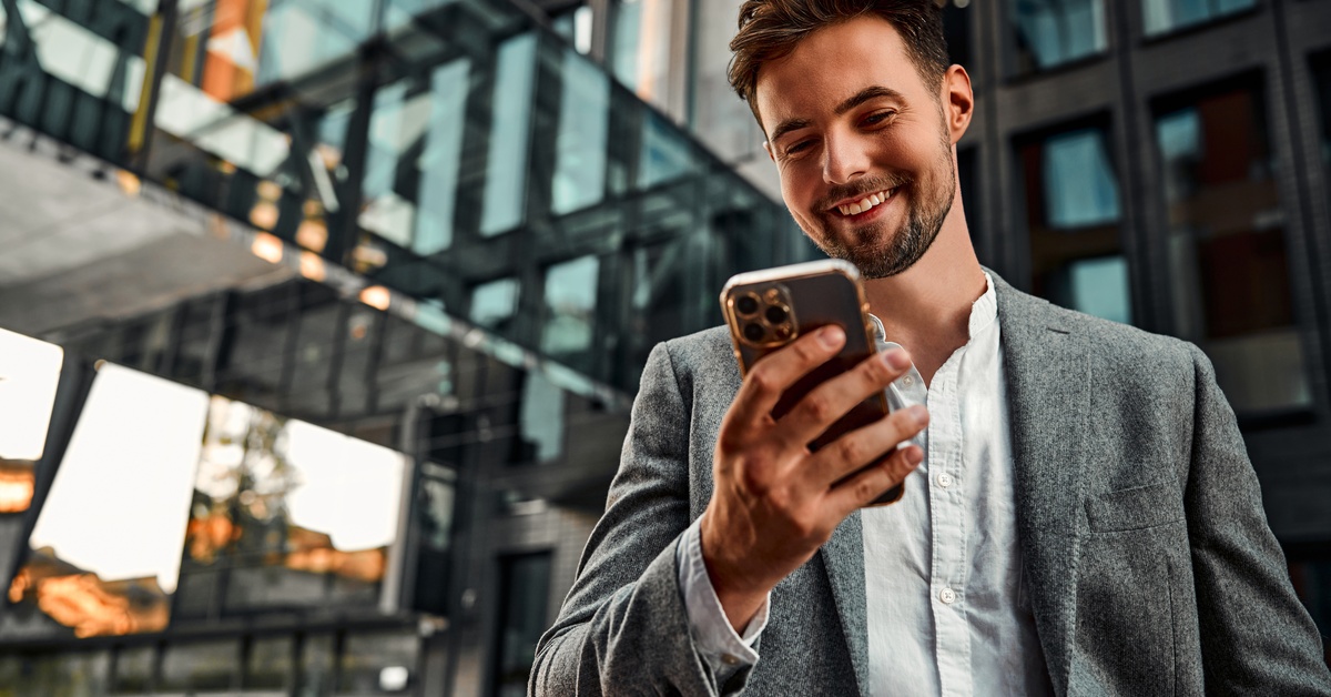 A man in business clothing stands outside in front of an office. He smiles at the smartphone in his hand.