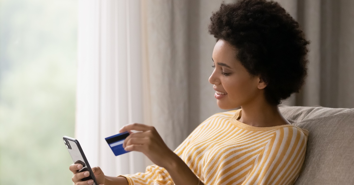 A woman sitting in an armchair holds her black smartphone in one hand and a credit card in the other.