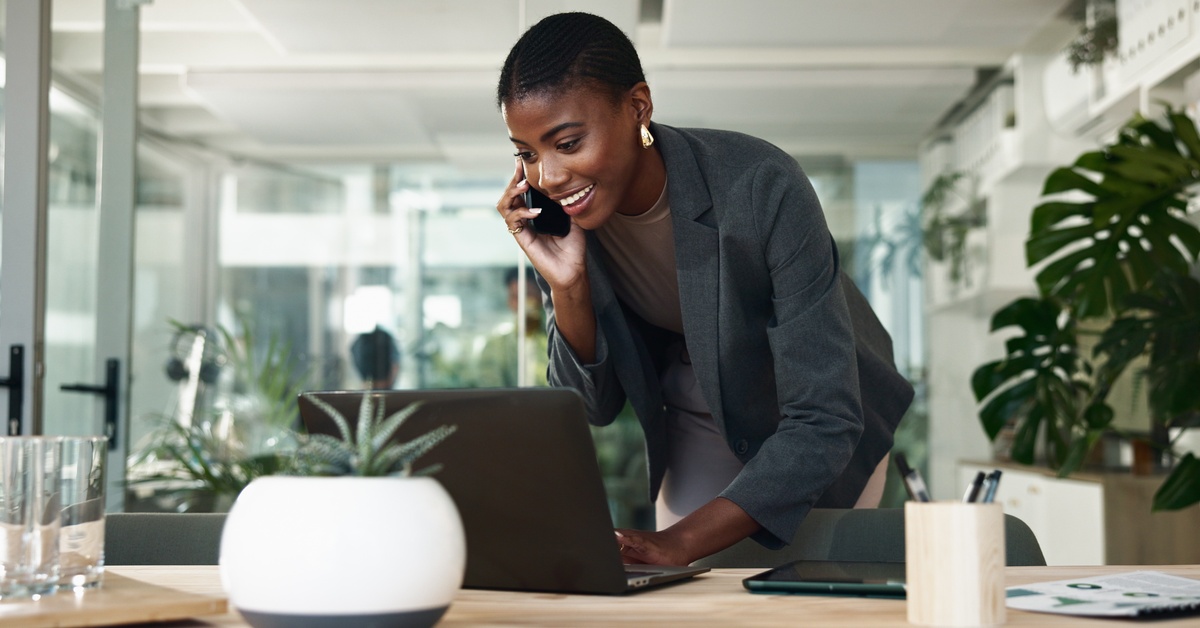 A woman in business clothing holds a phone to her ear as she leans over a desk and types at a laptop.