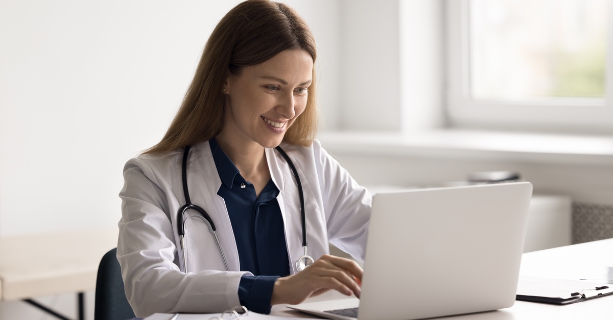 A medical professional wearing a white coat and stethoscope sits at a desk with a laptop open in front of her.
