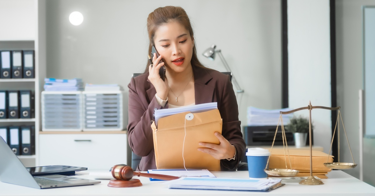 A woman sits at a desk and talks on the phone while holding papers. There is a gavel and scale of justice on the desk.
