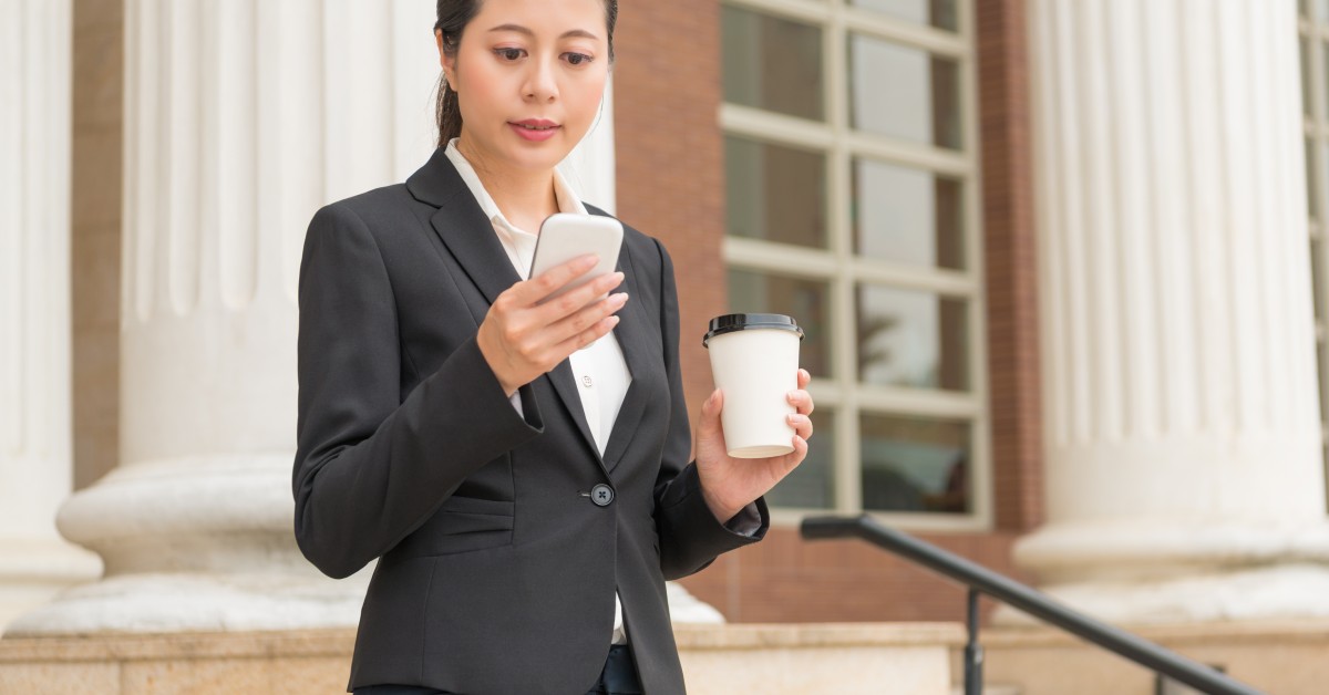 A woman in business clothing stands outside a building. She holds her phone and looks at it while holding a cup of coffee in the other hand.