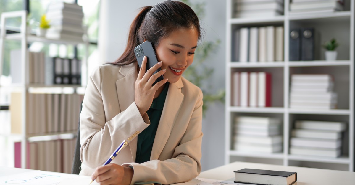 A woman sits at a desk with bookshelves in the background. She holds a pen and a phone to her ear while looking at paperwork on her desk