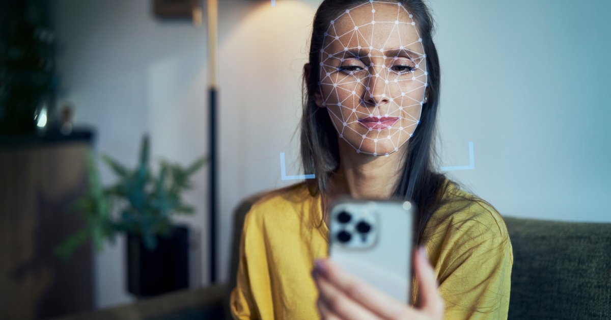 A woman sitting on her couch. She holds a phone in front of her with facial recognition graphics over her face.