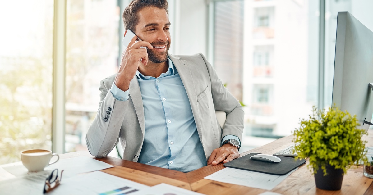 A businessman at his desk talking on the phone. His desk is cluttered with papers, glasses, coffee, and a plant.