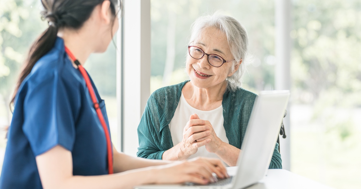 A healthcare worker sits at a table with an elderly patient. They look at each other as the worker types on her laptop.