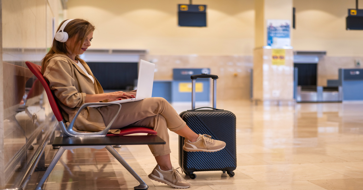 A woman sits in a chair in an airport. She has headphones on with a laptop resting on her lap and luggage next to her.