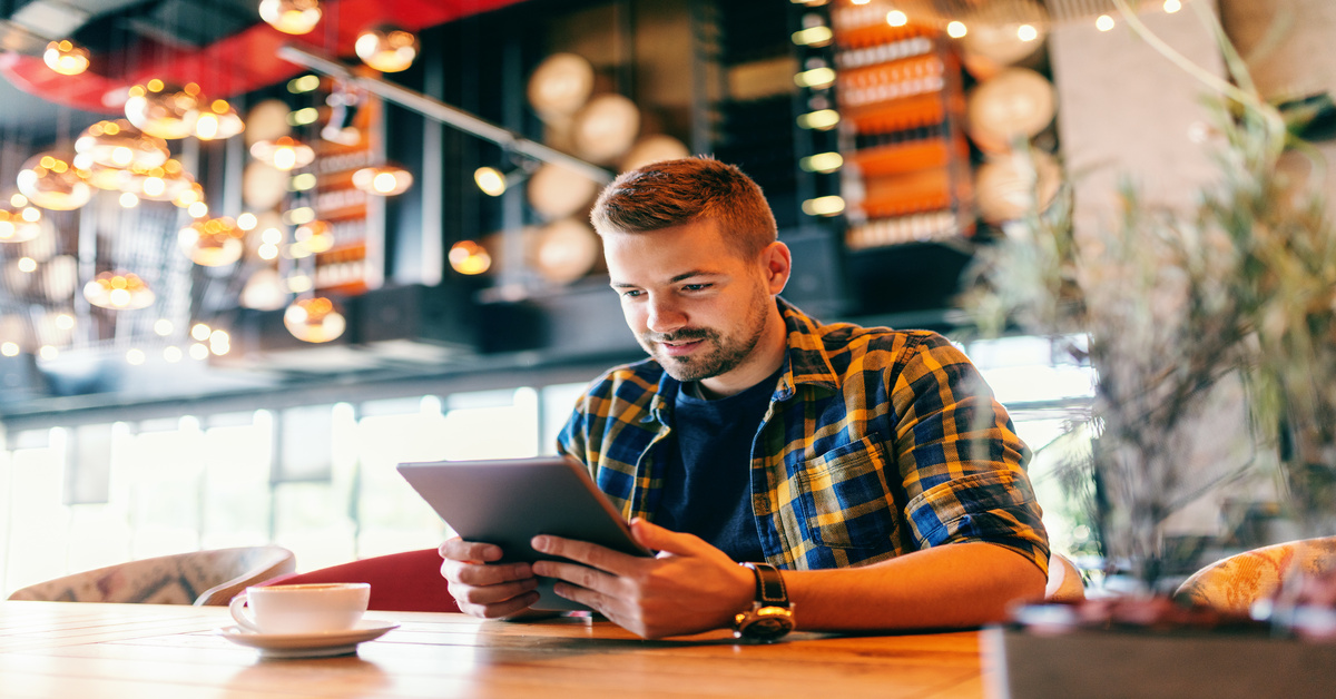 A man sits at a table in a restaurant. He has a cup of coffee, and he holds a tablet in front of him.