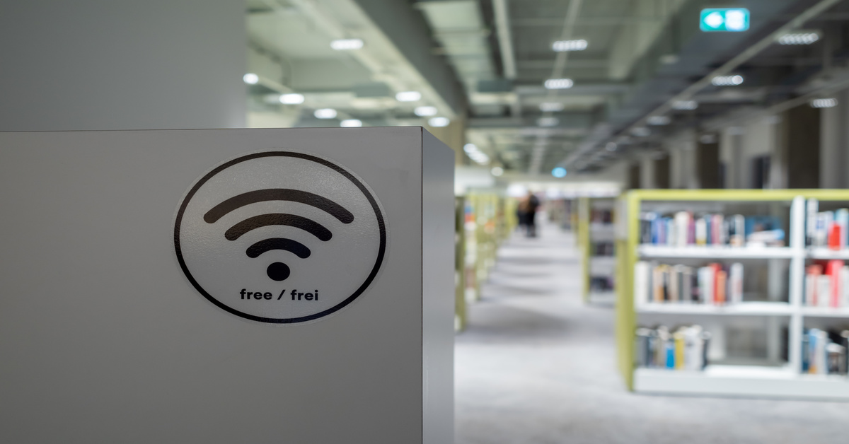 A close-up of a sign with a Wi-Fi symbol with the words "free/frei." There are bookshelves filled with books behind the sign.