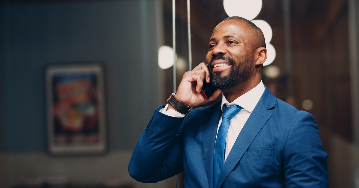 A man in a navy suit stands in an office with bright lighting behind him. He is smiling with a phone to his ear.