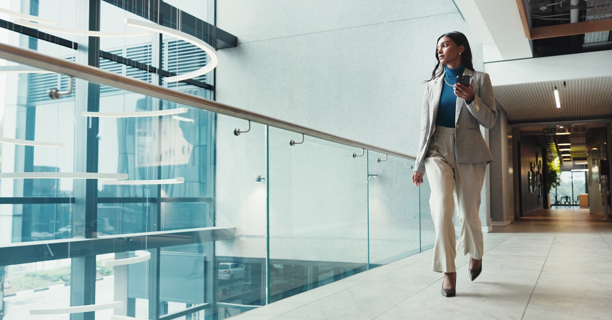 A woman in a cream-colored pant suit walks across a skywalk in an office. She holds a cell phone in her hand.