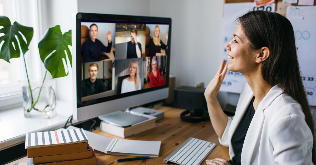 A woman sits at a desk with her hand waving toward the computer screen. The screen shows other people waving on a video call.