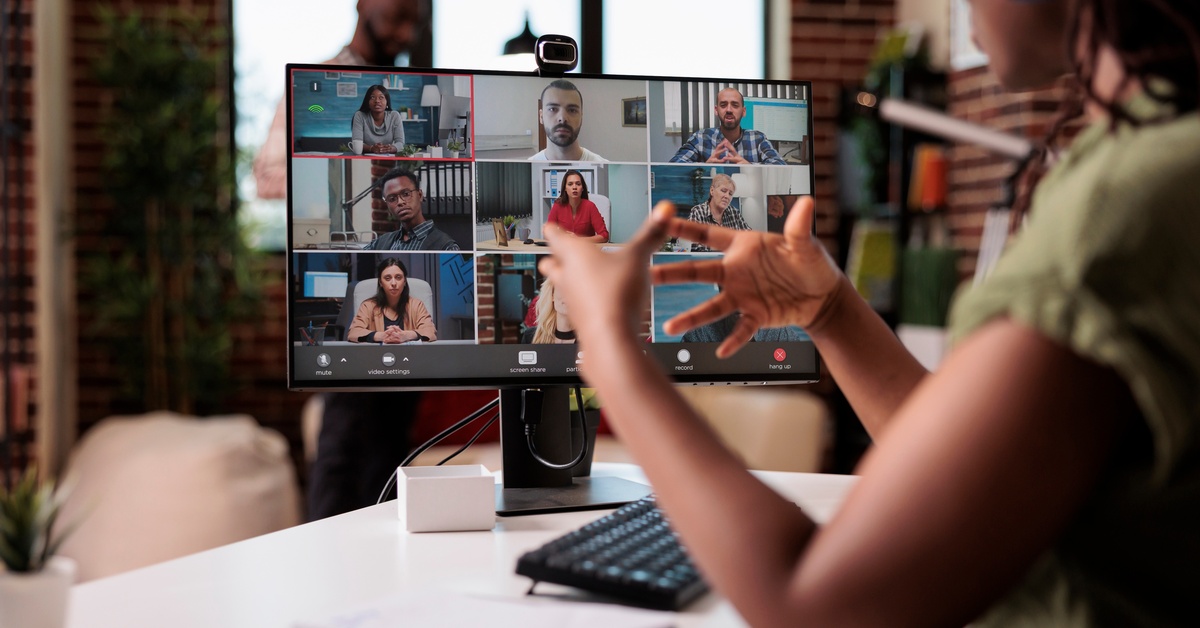 A woman sits at a desk and uses her hands to explain something. More coworkers are watching on a video call.