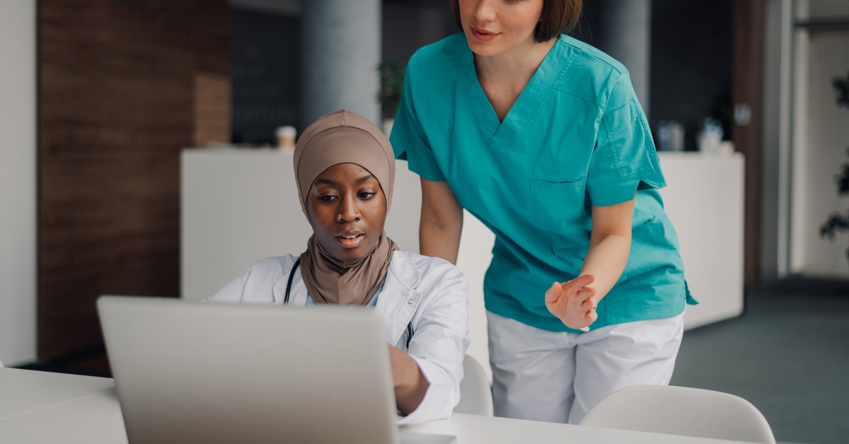 A doctor sitting at a table with a laptop open in front of her. A nurse stands behind her, looking at the screen.