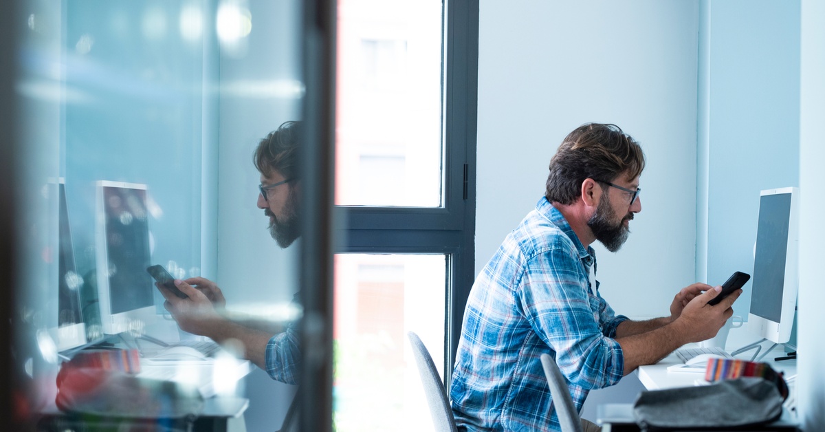 A man in a flannel shirt sits at a desk with a computer in an office space and looks at his cell phone.